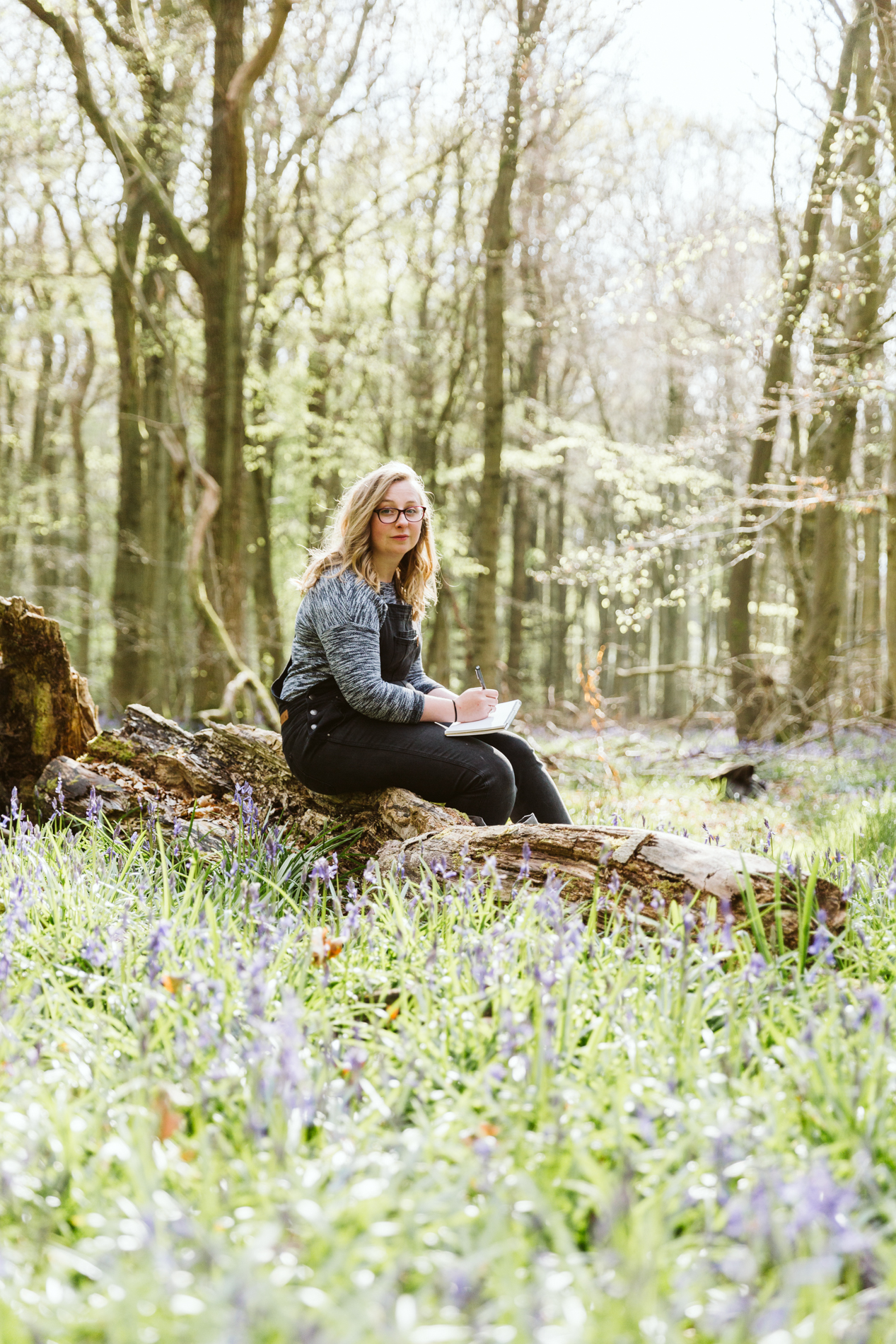 Gemma sat on tree sketching in woodland full of a bluebells 